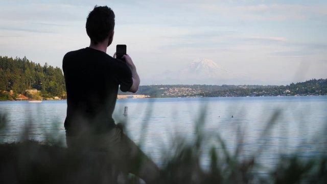 Static shot of young man taking pictures and selfies with a cell phone in front of Mt. Rainier at Seward Park in Seattle, Washington at dusk.