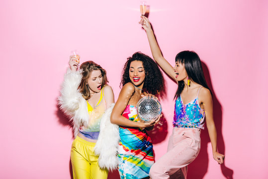 excited multicultural girlfriends with champagne and disco ball having fun on pink