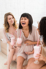 Obraz na płótnie Canvas cheerful multicultural girlfriends and laughing pregnant woman holding glasses with milkshakes on baby shower