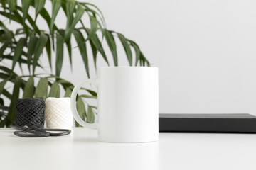Mug mockup with workspace accessories on a white table and a palm plant.