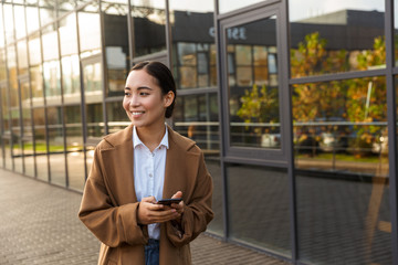 Image of young brunette asian woman using mobile phone on city street