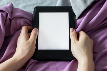 A modern black e-reader electronic book with a blank screen in female hands on a gray and purple bed. Mockup tablet on microfiber bedding closeup