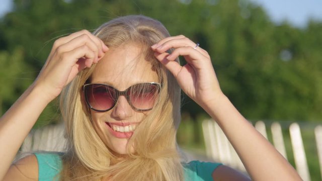 SLOW MOTION, CLOSE UP, PORTRAIT, DOF: Young woman laughs while playing with her gorgeous long blonde hair. Playful Caucasian girl runs her hand through her hair while having fun in the idyllic park.
