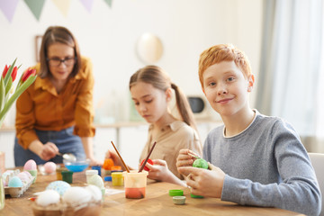 Red haired boy smiling at camera while sitting at the table and painting eggs together with his family