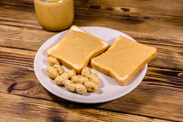 Glass jar with peanut butter and plate with sandwiches on a wooden table