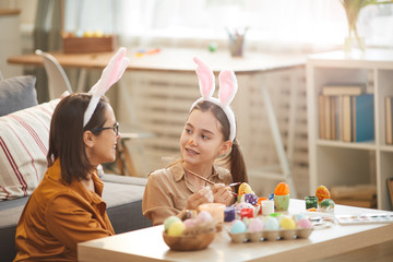 Little girl in rabbit ears sitting at the table and painting Easter eggs together with her mother at home