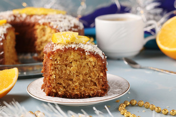 Homemade muffin with oranges sprinkled with coconut flakes on a light blue background and a cup of coffee, a piece of muffin in the foreground
