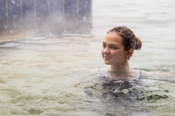 Girl in the pool with warm mineral water from a thermal spring in winter in snowy weather outdoors. Steam rises from warm water.
