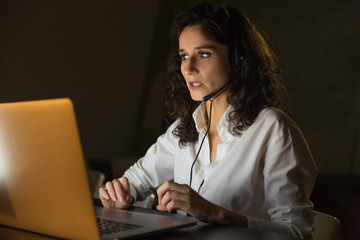 Businesswoman in headset working in dark office. Serious young businesswoman using headset and laptop computer while working late at night. Client support concept