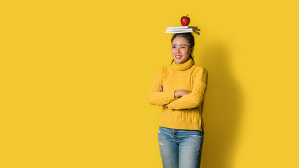 Cheerful young woman on yellow background in studio with a book and a red apple on her head. A...