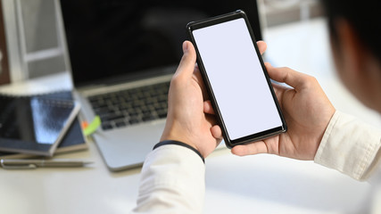 Cropped shot of businessman's hand holding crop black smartphone with white blank screen at the modern working desk with blurred laptop and office equipment as background.