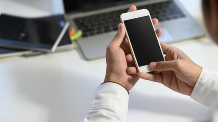 Photo of young businessman's hand holding white smartphone with black blank screen at modern working table with blurred laptop and office equipment as background.