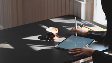 Cropped shot of young executive woman drawing on the computer tablet with white blank screen while planning about her business and sitting at the modern working desk. with office room as background.