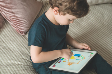 Boy sitting with a tablet in the room. The boy lies on a bed and plays on the tablet.