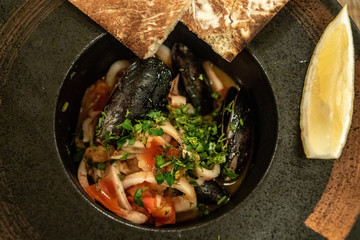 Roast mussels and squid with tomatoes and herbs in a dark plate with lemon and slices of bread. Delicacy