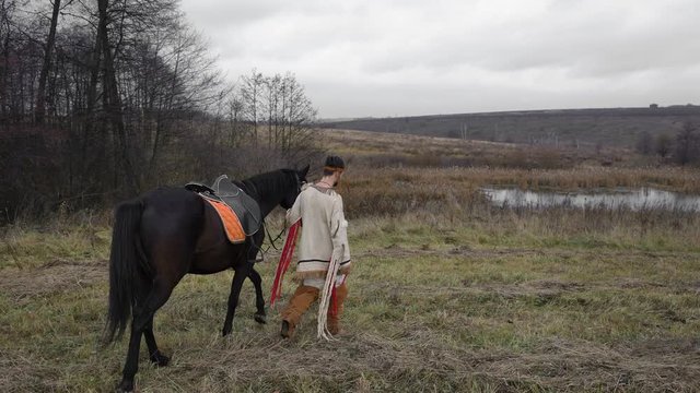 A young man in a white, beige suit with red threads Native American, leads a black beautiful horse along the forest to the lake in cloudy weather in autumn and turns to the camera