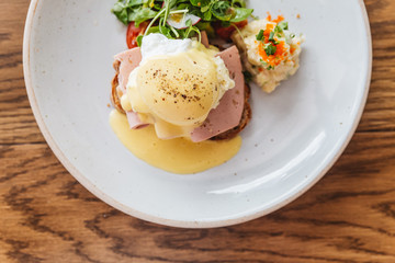 Top view of Eggs Benedict with ham, toast and mashed potato. Served with salad on a white plate on wooden table.