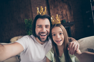 Closeup photo of little funny lady her handsome daddy friendship sit comfy sofa making taking selfies hugging wear golden crowns celebrities famous person house room indoors