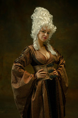 Portrait of medieval young woman in brown vintage clothing standing on dark background. Female...