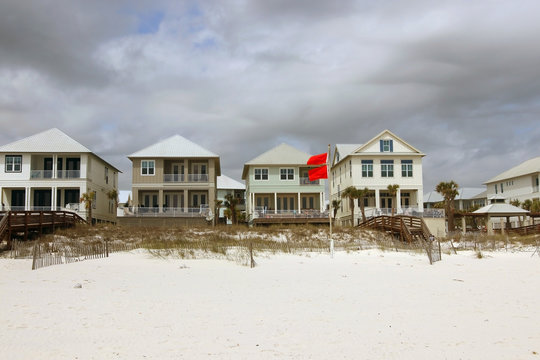 Row of oceanfront houses for vacation rentals behind sand dunes and two red flags symbolizing extremely danger and prohibition of swimming at Alabama Gulf Shores beach area, USA.