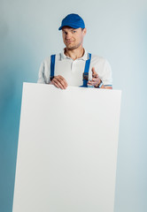 Mockup image of young smiling worker man wearing blue uniform. Holding white empty banner in his hand and pointing finger at you looking at camera. Isolated on grey background with copy space. Busines