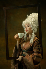 Drinking coffee. Portrait of medieval young woman in vintage clothing with wooden frame on dark...