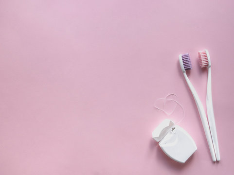on a pink background, two toothbrushes with white handles and pink and purple bristles hug. next lies dental floss in the form of a heart copy space
