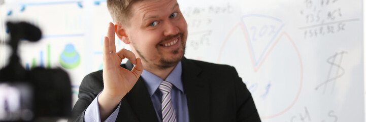 Male in suit and tie show confirm sign arm making promo videoblog or photo session in office camcorder to tripod portrait. Vlogger promotion selfie solution or finance advisor management information © H_Ko