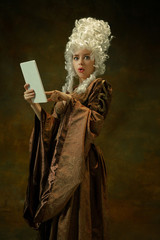 Obraz premium Astonished using tablet. Portrait of medieval young woman in brown vintage clothing on dark background. Female model as a duchess, royal person. Concept of comparison of eras, modern, fashion, beauty.