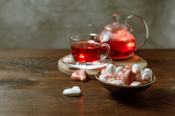 Beautiful romantic breakfast with red tea Hibiscus and meringue cookies in the shape of hearts. Glass tea-set stand on a dark wooden table with a concrete background. Still life with copy space.
