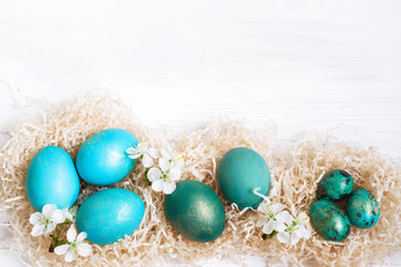 Fototapeta na wymiar Blue and green easter eggs in decorative nest with white spring flowers on a white wooden background closeup, top view. Easter background. 