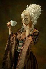 Drinking coffee with mask. Portrait of medieval young woman in brown vintage clothing on dark...