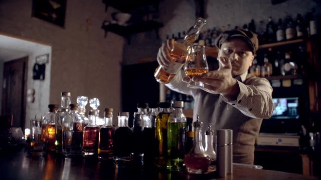 The bartender pours whiskey into a tasting glass (glencairn).  A bar counter in the speakeasy style. Dark scene, slow motion, camera movement.