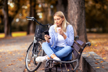 Fototapeta na wymiar Blonde young woman sitting on the bench looking on mobile phone and drinking coffee to go in park with bicycle on the background
