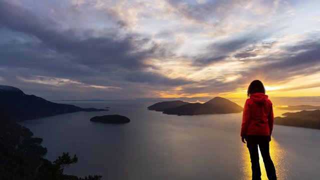 Time Lapse. Adventure Female Hiker on top of a high rocky mountain peak during a vibrant winter sunset. Composite Image Animation. Taken in Howe Sound near Vancouver, British Columbia, Canada.