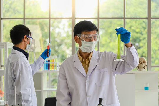 Portrait of senior asian doctor wearing medical particulate protective mask, laboratory glasses and blue rubber gloves holding laboratory glassware tube beaker with green liquid substance in researc