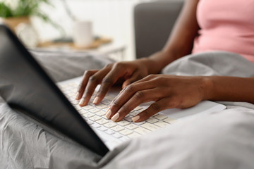 African-American woman using laptop computer in the bed in the morning. Everyday use of technology concept. Close-up detail.