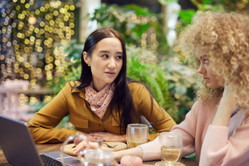 Young Asian woman discussing some project with her colleague while they sitting at the table with laptop in cafe