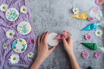 Homemade gingerbread cookies in the shape of flowers. Process of decorating spring gingerbread cookies (flowers) with colored icing with woman hands and spring flowers. Flat lay. Copy space.