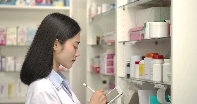 Young asian pharmacist woman holding computer tablet using for checking medication details on a box in pharmacy drugstore.