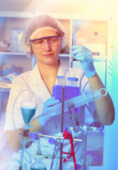 Young female scientist or tech in protective wear supervises chemical synthesis, checking reaction. Laboratory, glassware, futuristic neon light, research facility out of focus.