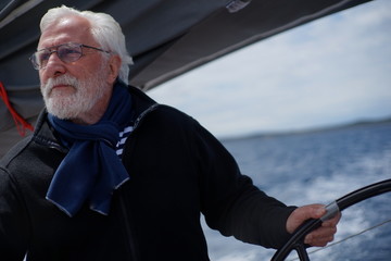White bearded sailing senior, experienced skipper on a sailboat holding the steering wheel while...