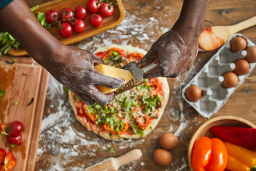 Close-up of man adding cheese on his homemade pizza while cooking at the wooden table in the kitchen