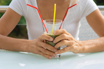 Female hands hold a glass with a green wholesome smoothie cocktail with a straw. Healthy raw breakfast of their vegetables in a country house