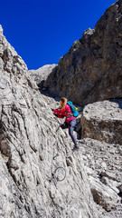 A woman with big hiking backpack climbing on a very steep via ferrata to reach the Grosse Gamswiesenspitze in Lienz Dolomites, Austria. Sharp and barren slopes. Massive Alpine mountains. Solo wanderer