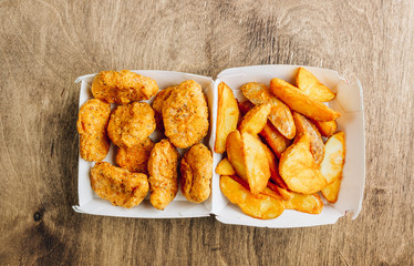 Serving in a paper box of takeaway crumbed fried nuggets with fried potato and a small bowl of sauce or dip on a rustic wooden background. Top view. Copy space