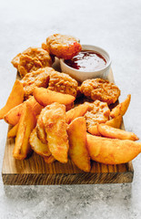 Serving on a wooden board crumbed fried nuggets with fried potato and a small bowl of sauce or dip on a stone background. Top view. Copy space