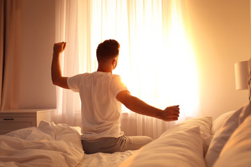 Young man stretching on bed at home, view from back. Lazy morning