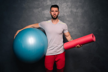 Portrait of attractive  muscular caucasian man holding a medical ball and a training mat, promoting a healthy lifestyle and fitness training