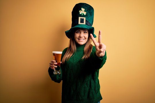 Young beautiful woman wearing green hat drinking glass of beer on saint patricks day smiling with happy face winking at the camera doing victory sign with fingers. Number two.